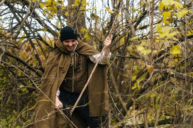 Portrait of smiling tourist male wearing raincoat tent chopping small branches with small shovel in forest on overcast cold day