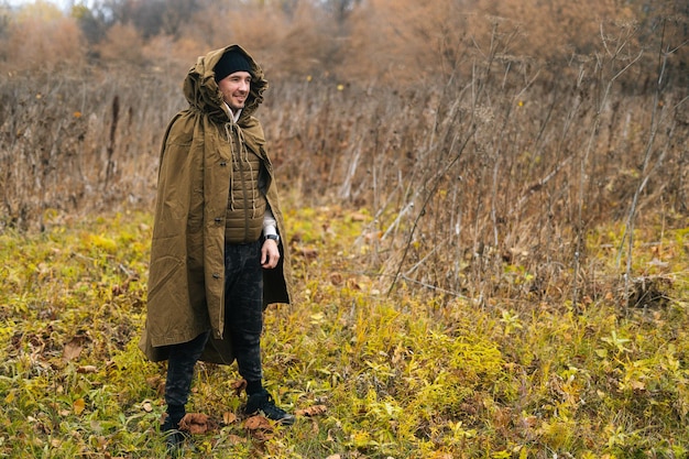 Portrait of smiling tourist man wearing green raincoat tent standing in thicket of bushes in cold overcast day and looking away