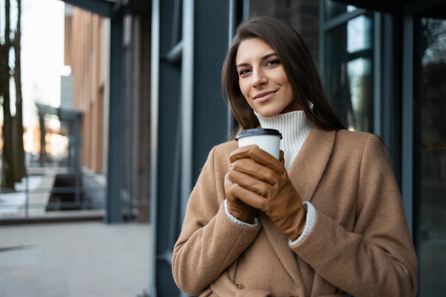 Portrait of smiling woman in coat with cup of coffee near the office building