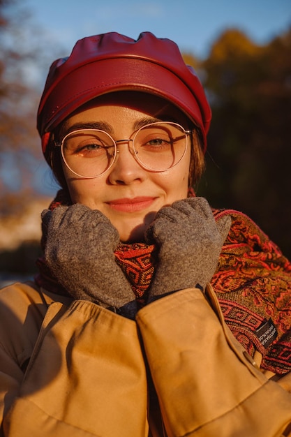 Portrait of smiling woman in glasses cap and gloves warming in scarf in autumn park