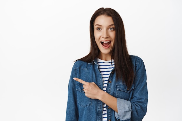 Portrait of smiling woman looks in awe, thrilled with promotion, pointing finger and looking left at awesome product, advertisement or logo, standing against white background.