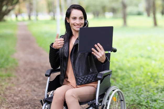 Portrait of smiling woman sitting in wheelchair in park and showing thumb up on tablet of