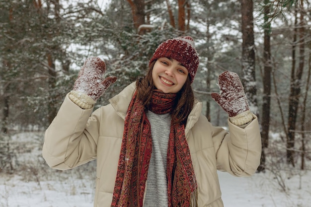 Portrait of smiling woman in warm outerwear and mittens in snow standing in winter forest