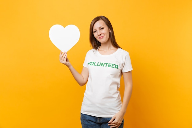 Portrait of smiling woman in white t-shirt with written inscription green title volunteer hold white heart isolated on yellow background. Voluntary free assistance help, charity grace work concept.