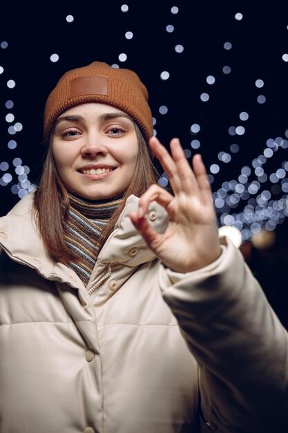 Portrait of smiling woman in winter outerwear showing okay gesture and looking at camera