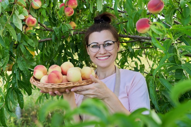 Portrait of smiling woman with basket of fresh peaches tree with ripe peaches background