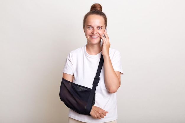 Portrait of smiling woman with bun hairstyle wearing casual Tshirt standing in arm sling isolated on light gray background talking via smart phone expressing happiness