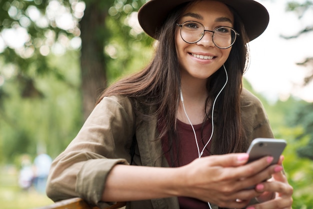 Portrait of smiling woman with long dark hair wearing hat and eyeglasses using earphones and smartphone in green park