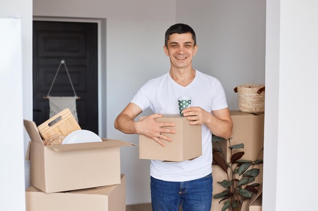 Portrait of smiling young adult Caucasian man wearing white t shirt standing with cardboard box and cup with coffee looking at camera with positive facial expression