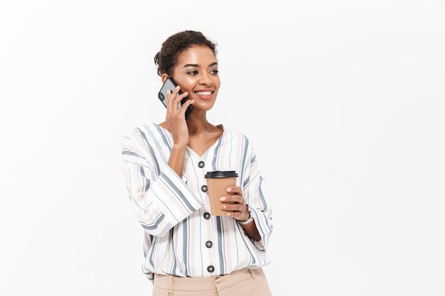 Portrait of a smiling young african businesswoman standing isolated over white wall, drinking takeaway coffee, talking on mobile phone