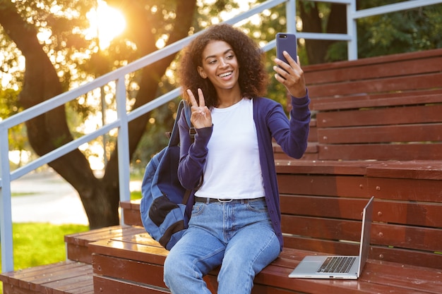 Portrait of a smiling young african girl with backpack resting at the park, taking a selfie