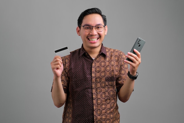 Portrait of smiling young Asian man wearing batik shirt holding credit card and smartphone isolated on grey background