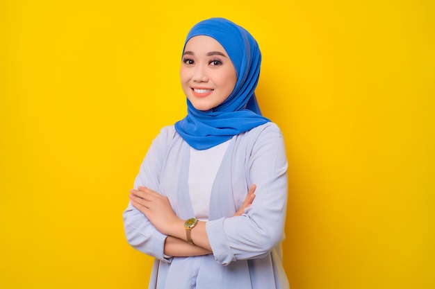 Portrait of smiling young Asian Muslim woman standing with crossed arms and looking at camera isolated over yellow background