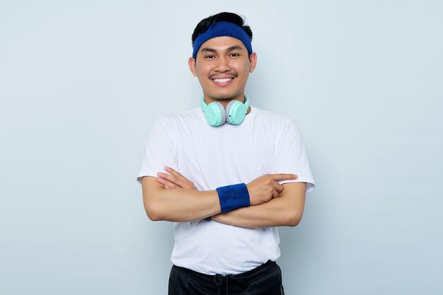 Portrait of Smiling young Asian sporty man in blue headband and white tshirt with headphonescrossed arms chest and looking confident isolated on white background Workout sport concept