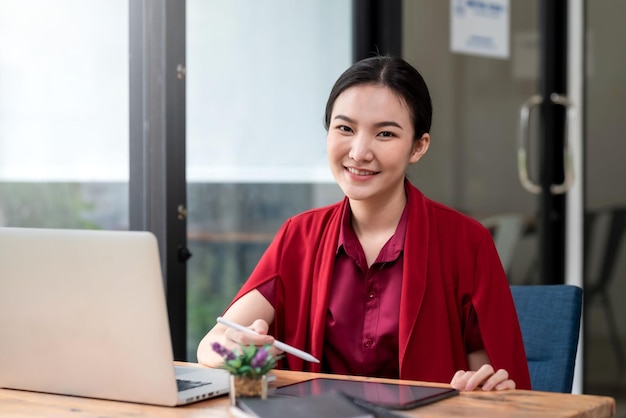 Portrait of smiling young beautiful Asian businesswoman sitting with laptop computer looking at camera in the office