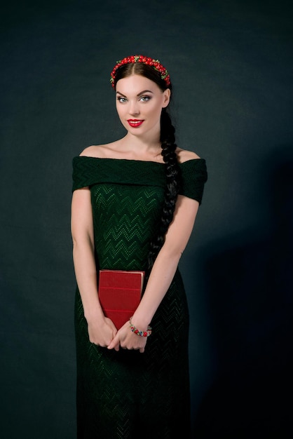 portrait of smiling young beautiful brunette with red wreath and braid on dark background