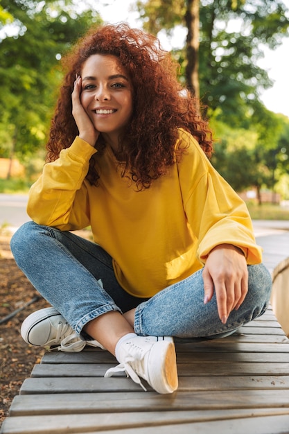 Portrait of a smiling young beautiful curly student outdoors in nature park sitting on a bench.