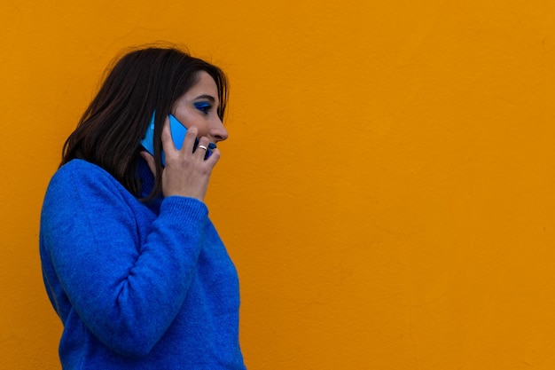 Portrait of smiling young brunette woman in blue dress and makeup talking on the phone on yellow background