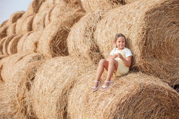 Portrait of smiling young girl wearing sundress sitting on tall haystack and leaning on another one and a lot of other haystacks in background Having fun away from city on field full of golden hay