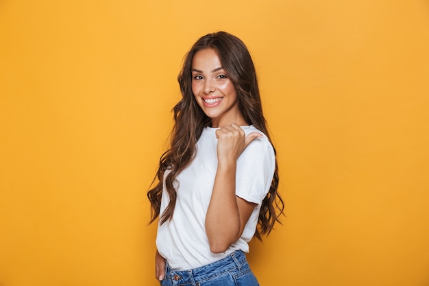 Portrait of a smiling young girl with long brunette hair standing over yellow wall, pointing away