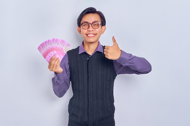 Portrait of smiling young handsome Asian businessman holding cash money in rupiah banknotes and showing thumb up isolated on white background People lifestyle concept