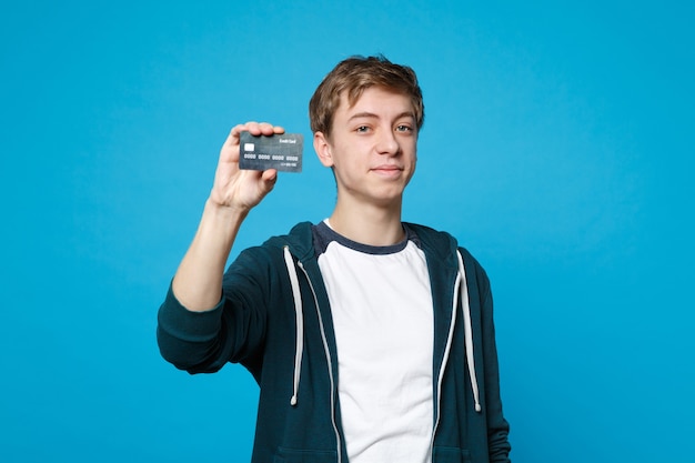 Portrait of smiling young man in casual clothes holding credit bank card isolated on blue wall . People sincere emotions, lifestyle concept. 