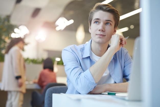 Portrait of smiling young man looking away dreamily while working with laptop in office