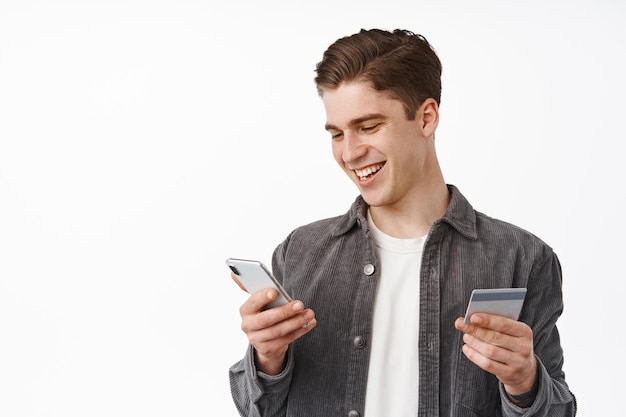 Portrait of smiling young man purchasing in mobile phone store, paying with smartphone and credit card, order delivery on cellphone, standing against white background