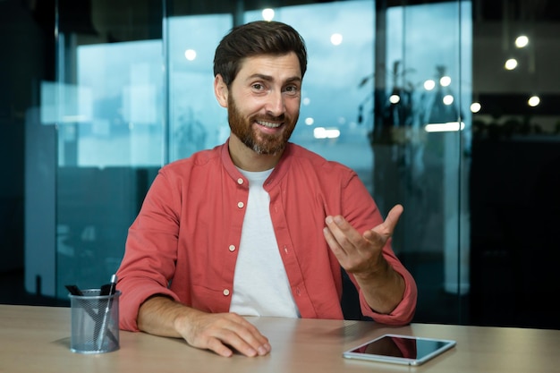 Portrait of a smiling young man sitting at the desk in the office in front of the camera and talking