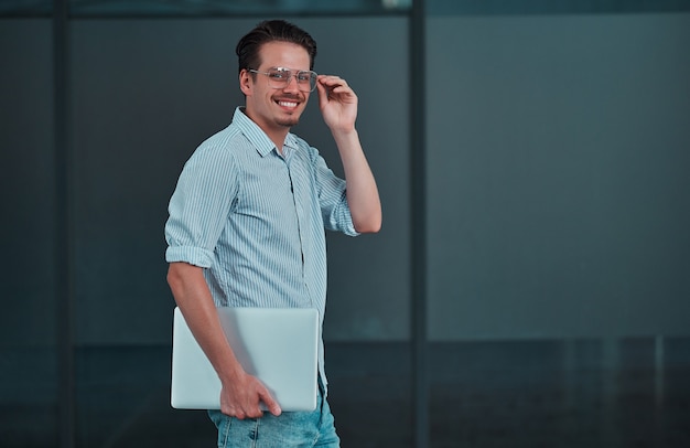 Portrait of a smiling young man standing with laptop against business center.