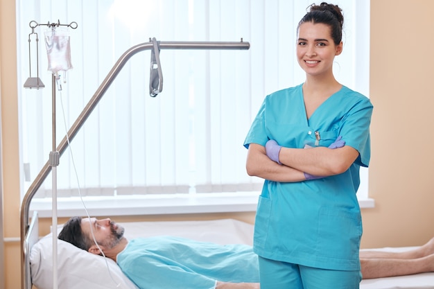 Portrait of smiling young nurse in surgical gloves standing with crossed arms at patients bed in hospital