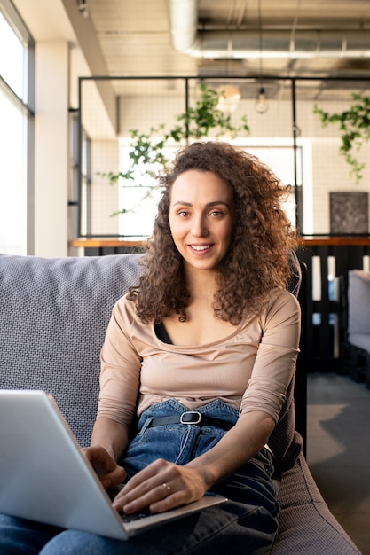 Portrait of smiling young SMM manager with curly hair sitting on comfortable sofa and working with laptop in cafe