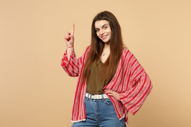 Portrait of smiling young woman in casual clothes looking camera pointing index finger up isolated on pastel beige background in studio. People sincere emotions, lifestyle concept. Mock up copy space.