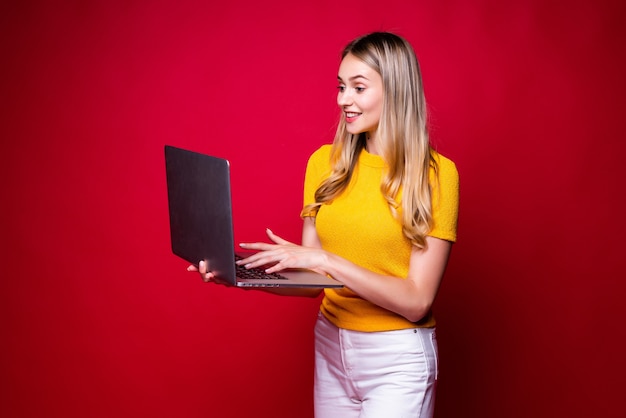 Portrait of smiling young woman holding, working on laptop pc computer isolated on red wall.