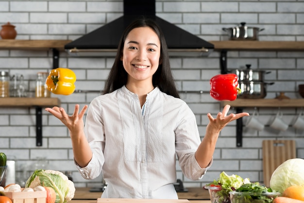 Portrait of smiling young woman juggling with bell peppers in kitchen