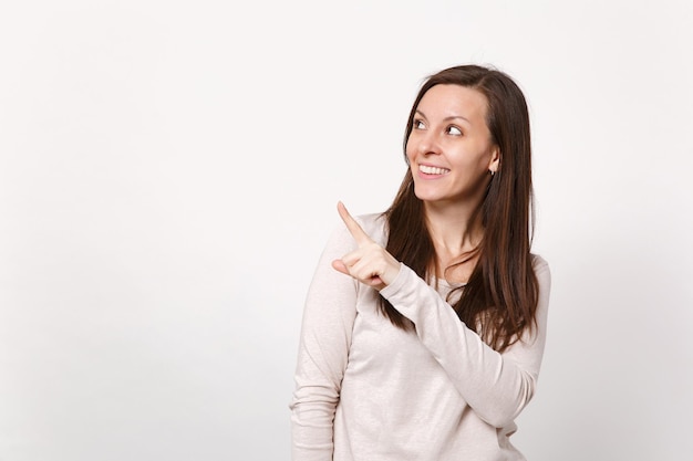 Portrait of smiling young woman in light clothes looking, pointing index finger aside up isolated on white wall background in studio. People sincere emotions, lifestyle concept. Mock up copy space.