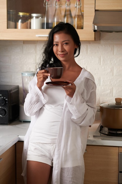 Portrait of smiling young woman in loungewear standing in kitchen and drinking cup of morning coffee