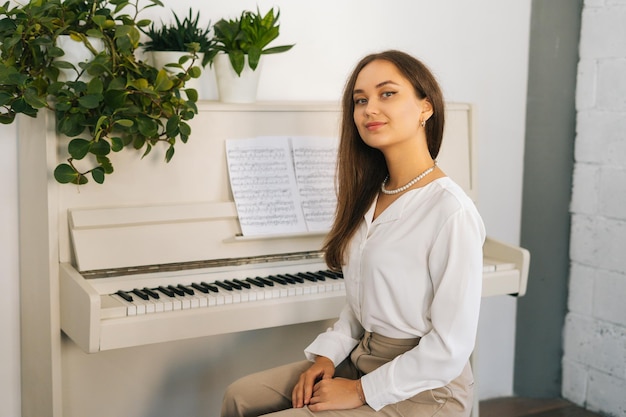 Portrait of smiling young woman pianist looking at camera sitting at classical white piano in classroom