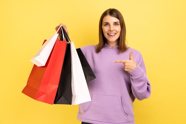 Portrait of smiling young woman pointing at shopping bags looking at camera with pleased happy facial expression wearing purple hoodie Indoor studio shot isolated on yellow background