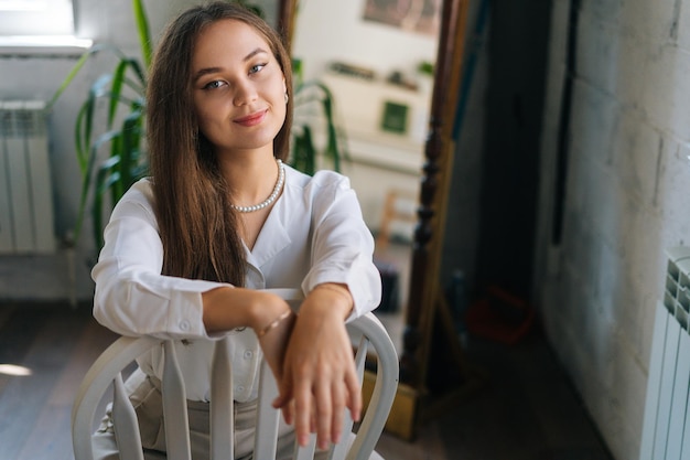 Portrait of smiling young woman sitting on chair with legs wide open and holding hands on back of chair looking at camera