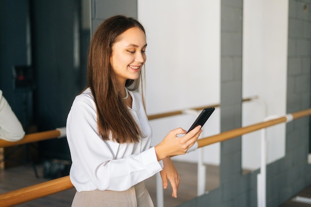 Portrait of smiling young woman in stylish clothes using browsing mobile phone standing in room with mirror looking on screen