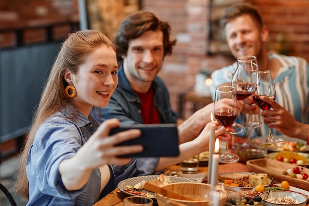 Portrait of smiling young woman taking selfie with friends at dinner table in cozy warm setting