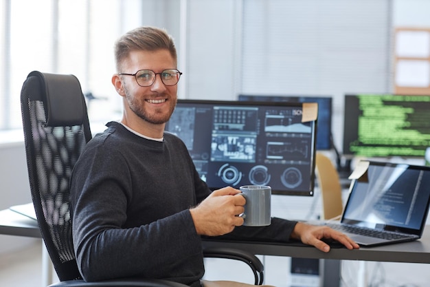 Portrait of software programmer smiling at camera while posing at workplace in office with coffee cu