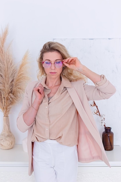 Portrait of stilysh woman in glasses and light clothes is standing in the white interior