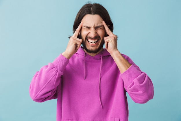 Portrait of a stressed young bearded brunette man wearing hoodie standing isolated over blue wall, suffering from a headache