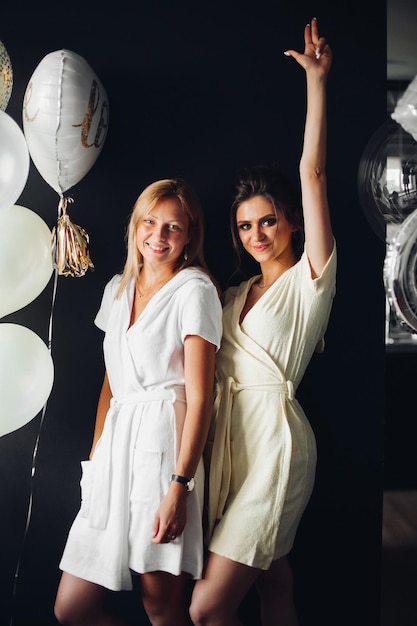 Photo portrait of strong and dangerous women with make up posing at camera gorgeous brunette girls holding their hands up and dancing happy friends chilling out at party near white and gold balloons