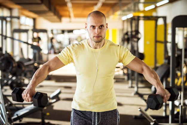 Portrait of strong muscular active healthy young man raising dumbbells with open arms and listening music in the gym.