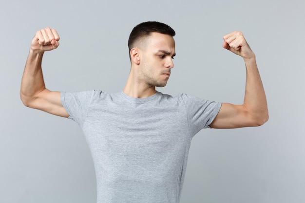 Portrait of strong young man in casual clothes spreading hands showing looking on biceps, muscles 