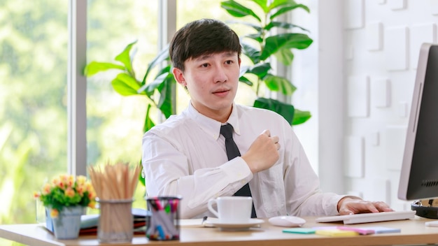 Portrait studio shot of Asian professional successful male businessman employee in formal shirt with necktie sitting look at camera at working desk with computer monitor keyboard mouse and stationery