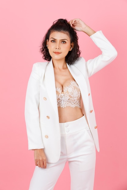 Portrait studio shot of Asian sexy curly hairstyle professional successful businesswoman in white fashionable casual suit with lace lingerie crop top bra standing look at camera on pink background